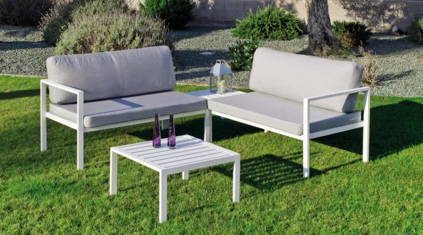 Rinconera de balcón colocada en un patio ajardinado.La rinconera esa pintada en color blanco y los cojines son de color gris.